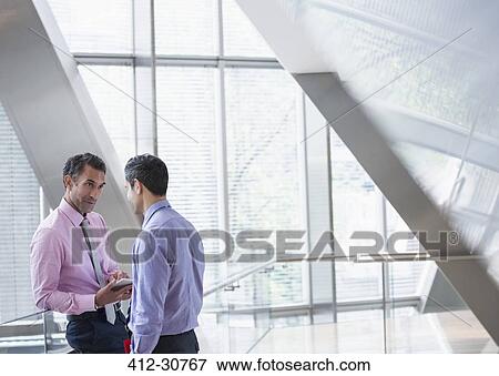 Stock Photo - Corporate businessmen with digital tablet talking in modern office lobby. Fotosearch
