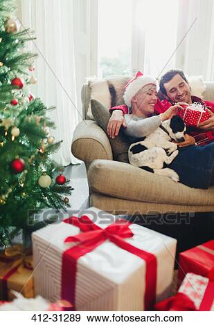 Stock Photo - Couple cuddling on sofa with dog near Christmas tree and gifts. Fotosearch