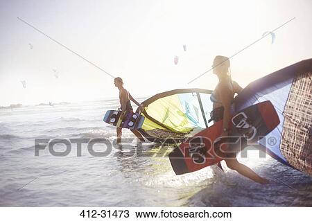 Stock Image - Couple walking into surf with kiteboard equipment. Fotosearch