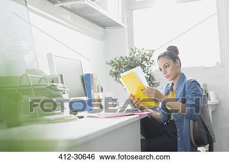 Stock Photograph - Creative young businesswoman reading paperwork at desk in sunny office. Fotosearch