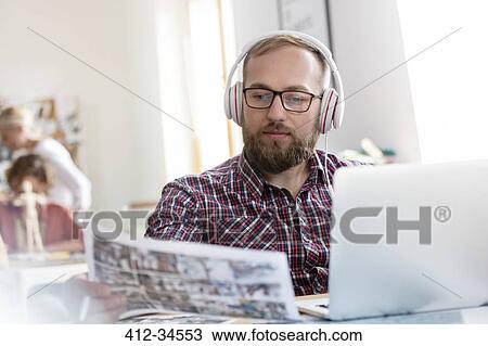 Stock Image - Design professional listening to music with headphones reviewing proofs at laptop. Fotosearch
