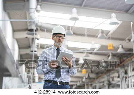 Engineer with hard-hat and digital tablet walking in factory View Large Photo Image Picture - Engineer with hard-hat and digital tablet walking in factory. Fotosearch