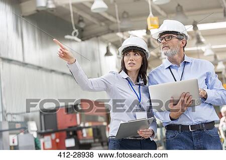 Stock Photo - Engineers with laptop and clipboard pointing in factory. Fotosearch