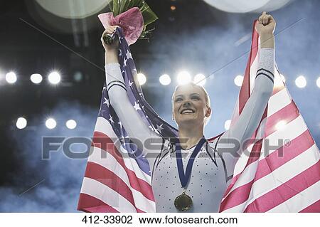 Stock Image - Enthusiastic female gymnast celebrating victory holding American flag on winners podium. Fotosearch