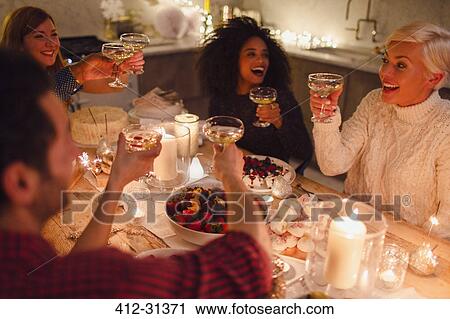 Stock Image - Enthusiastic friends toasting champagne glasses at candlelight Christmas dinner. Fotosearch