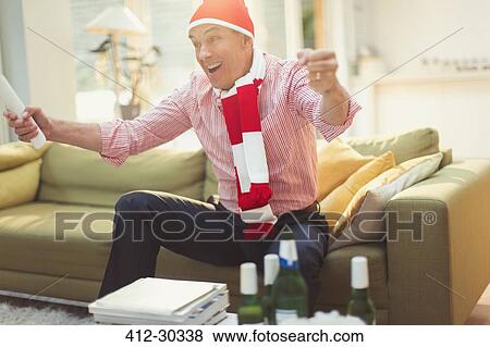 Enthusiastic mature man in hat and scarf watching TV sports event on living room sofa View Large Photo Image Stock Photo - Enthusiastic mature man in hat and scarf watching TV sports event on living room sofa. Fotosearch