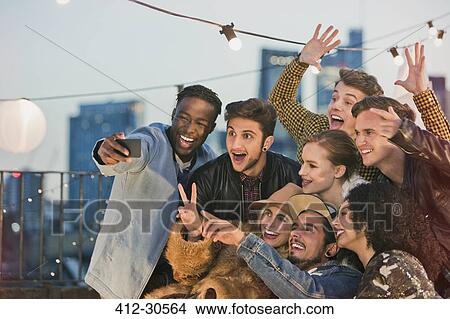 Picture - Enthusiastic young adult friends cheering and taking selfie at rooftop party. Fotosearch
