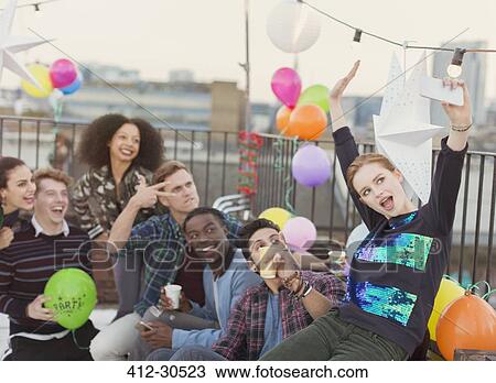 Stock Image - Enthusiastic young adult friends taking selfie at rooftop party. Fotosearch