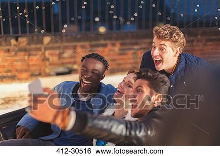Stock Photograph - Enthusiastic young adults taking selfie at nighttime rooftop party. Fotosearch