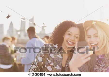 Picture - Enthusiastic young women taking selfie at rooftop party. Fotosearch