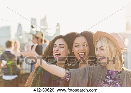 Stock Image - Enthusiastic young women taking selfie at rooftop party. Fotosearch