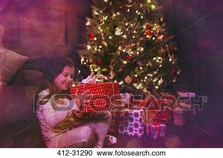 Stock Image - Excited girl holding gift next to Christmas tree. Fotosearch