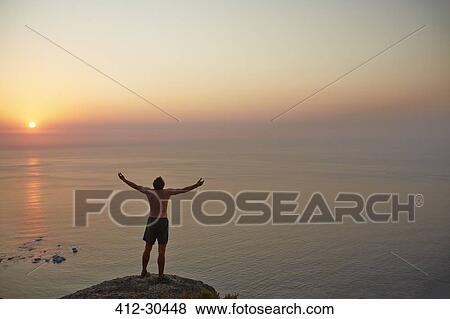 Exuberant male runner with arms outstretched on rock overlooking ocean sunset View Large Photo Image Stock Photo - Exuberant male runner with arms outstretched on rock overlooking ocean sunset. Fotosearch