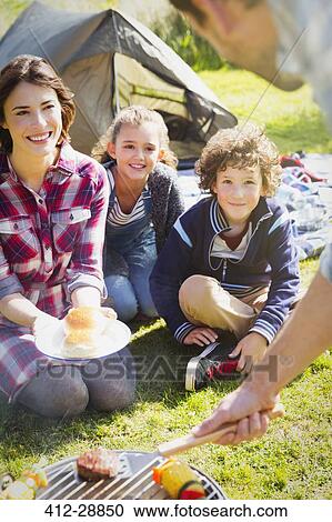 Stock Image - Family watching father barbecuing at campsite grill. Fotosearch