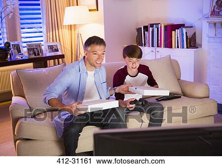 Stock Image - Father and son eating pizza and playing video game in living room. Fotosearch