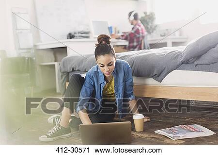 Stock Image - Female college student with coffee using laptop on bedroom floor. Fotosearch