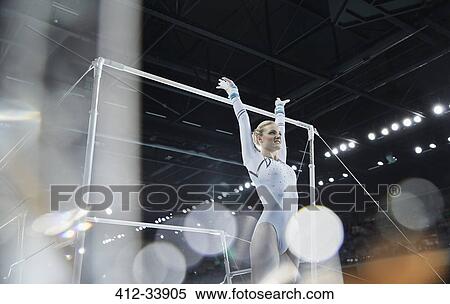 Stock Photography - Female gymnast with arms raised below uneven bars in arena. Fotosearch