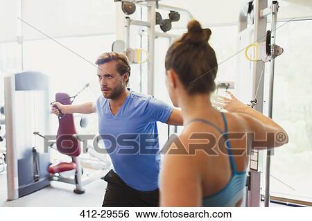 Stock Photograph - Female personal trainer guiding man doing cable chest fly at gym. Fotosearch
