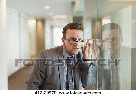 Stock Photo - Focused businessman eavesdropping with glass in office corridor. Fotosearch