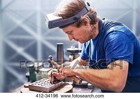 Stock Photograph - Focused jeweler using hammer in workshop. Fotosearch