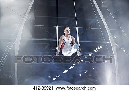 Stock Image - Focused male gymnast performing on gymnastics rings in arena. Fotosearch