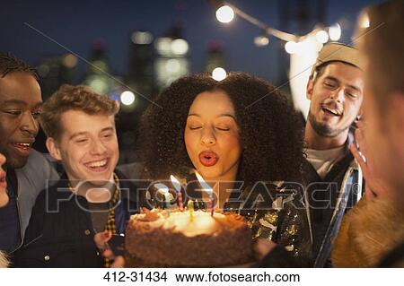 Picture - Friends cheering young woman blowing out birthday candles. Fotosearch