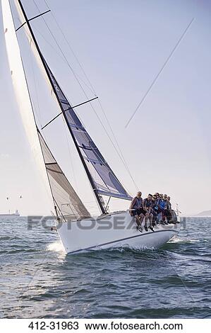 Stock Image - Friends sailing on heeling sailboat on ocean under blue sky. Fotosearch