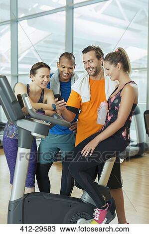 Stock Image - Friends using cell phone at exercise bike in gym. Fotosearch