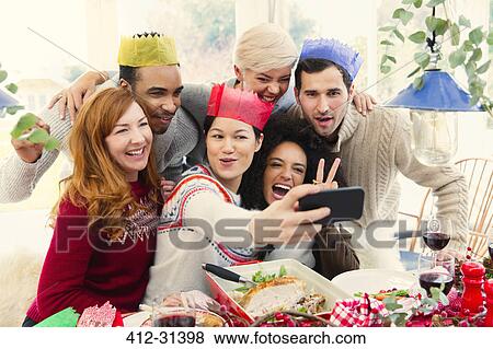 Stock Photo - Friends wearing paper crowns taking selfie at Christmas dinner. Fotosearch