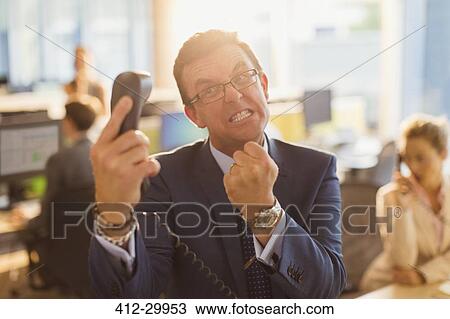 Stock Image - Furious businessman gesturing with fist at telephone in office. Fotosearch