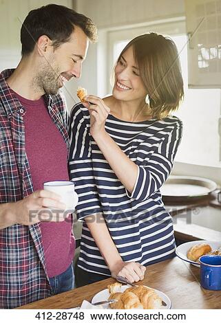 Stock Photo - Girlfriend feeding boyfriend croissant in kitchen . Fotosearch