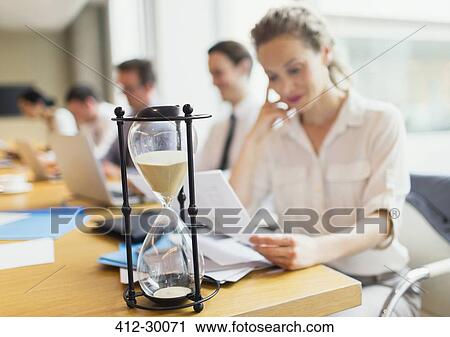 Stock Image - Hourglass in front of businesswoman reviewing paperwork in conference room. Fotosearch