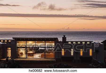 Stock Image - Illuminated luxury home overlooking ocean at sunset. Fotosearch