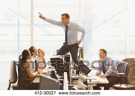 Stock Photo - Laughing colleagues watching exuberant businessman dancing on top of desks in office. Fotosearch