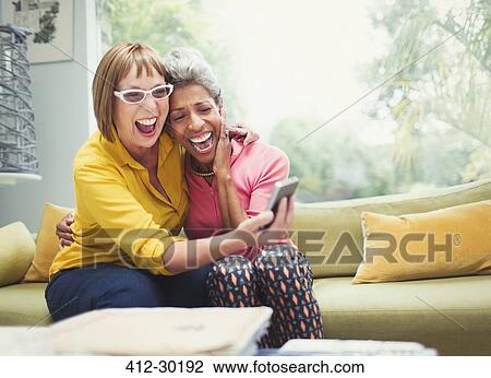 Stock Image - Laughing mature women hugging and taking selfie on sofa. Fotosearch