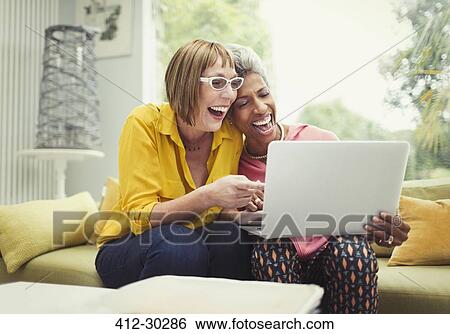Stock Photograph - Laughing mature women sharing laptop on living room sofa. Fotosearch