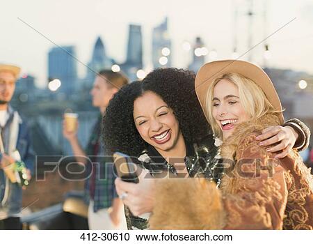 Laughing young women taking selfie at rooftop party View Large Photo Image Stock Image - Laughing young women taking selfie at rooftop party. Fotosearch