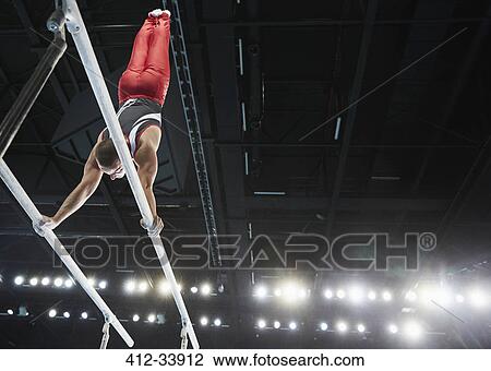 Stock Image - Male gymnast performing upside-down handstand on parallel bars in arena. Fotosearch