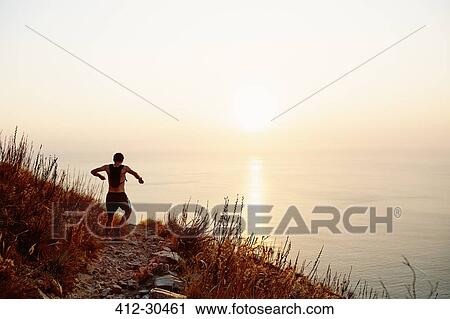 Stock Image - Male runner with backpack descending craggy trail overlooking sunset ocean. Fotosearch