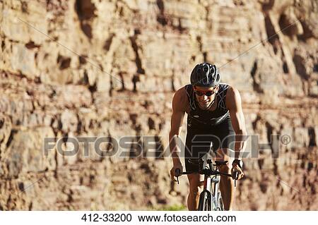 Stock Image - Male triathlete cyclist cycling along sunny rocks. Fotosearch