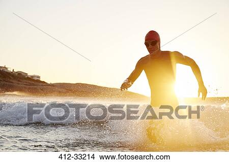 Picture - Male triathlete swimmer in wet suit running out of sunny ocean. Fotosearch