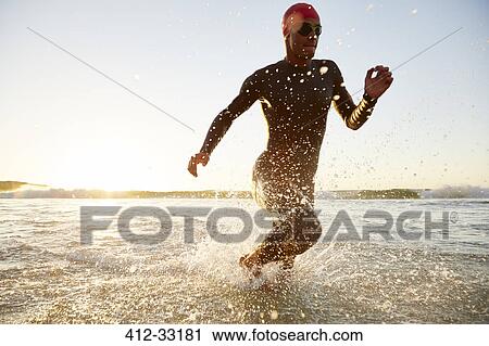 Stock Image - Male triathlete swimmer in wet suit running from ocean. Fotosearch