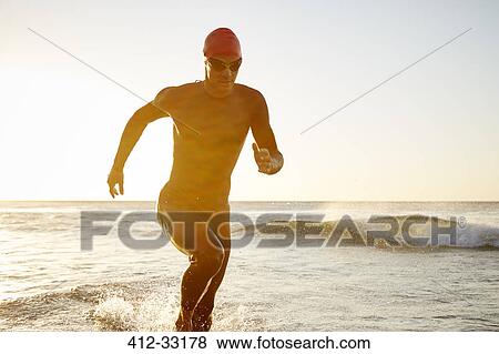 Stock Photo - Male triathlete swimmer in wet suit running from ocean. Fotosearch