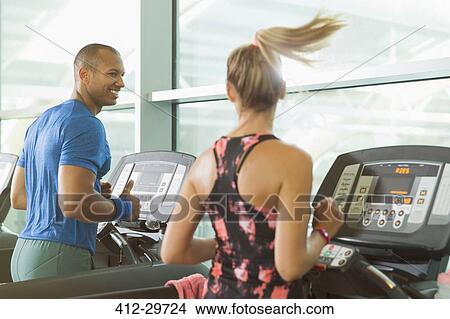 Picture - Man and woman talking and running on treadmills at gym. Fotosearch