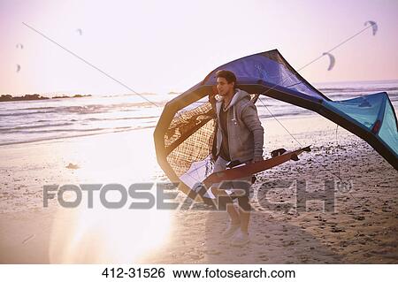 Stock Photograph - Man carrying kiteboarding equipment on sunset beach. Fotosearch