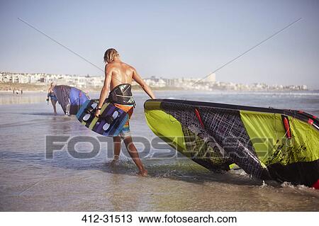 Stock Image - Man dragging kiteboarding equipment into ocean surf. Fotosearch