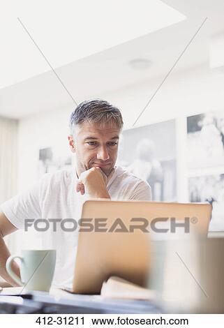 Stock Image - Man drinking coffee and working at laptop in kitchen. Fotosearch