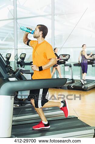 Stock Image - Man drinking water and running on treadmill at gym. Fotosearch