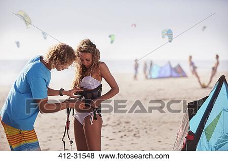 Stock Photo - Man fastening kiteboarding safety harness onto woman on sunny beach. Fotosearch