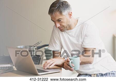 Stock Image - Man in pajamas drinking coffee and working at laptop in kitchen. Fotosearch